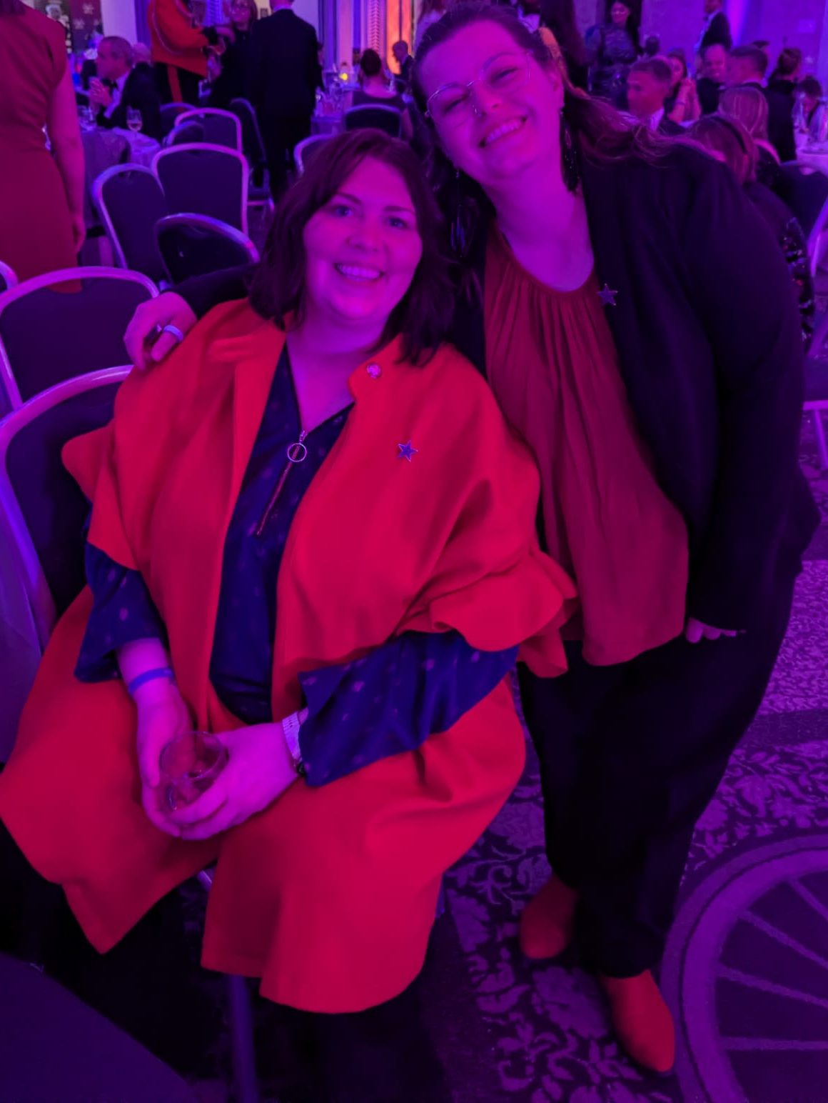 Two women smile together at a formal awards event in a large ballroom with purple lighting, chandeliers and guests seated at tables in the background.