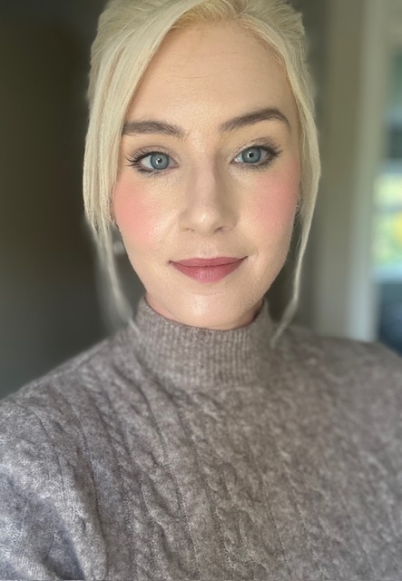 A woman with blonde hair wearing a grey knitted jumper looks directly at the camera in a close-up portrait photo.