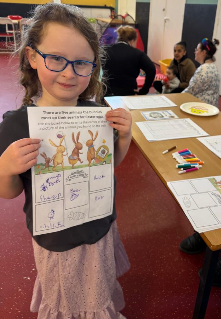 A child proudly holds up a completed Easter-themed worksheet during a community activity session. The worksheet shows drawings and writing about animals the bunnies meet on an Easter egg trail. Other people are seated at a table in the background with pens, worksheets and craft materials.