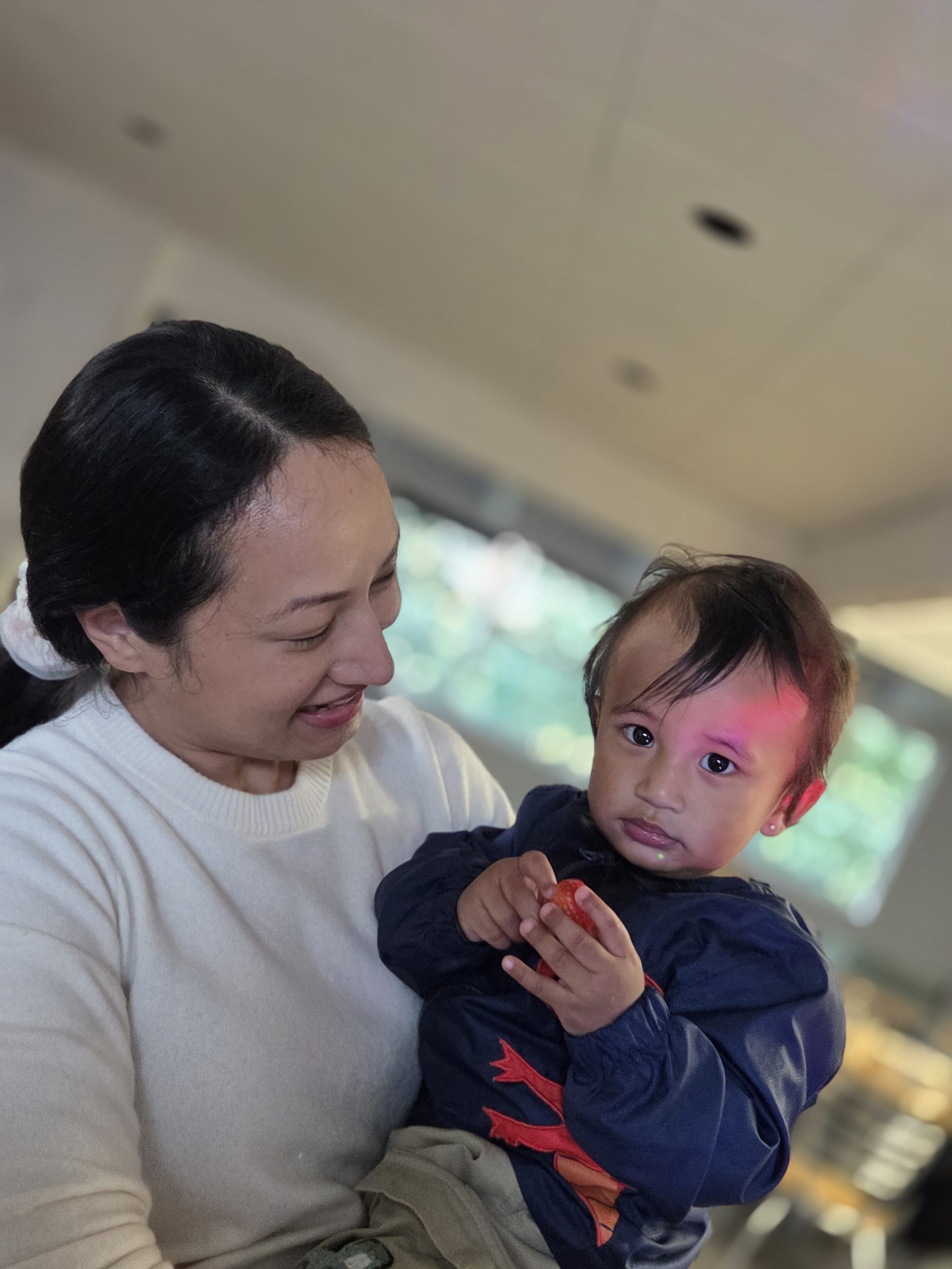 An adult smiling warmly while holding a young child indoors. The child is wearing a dark blue top with red detail and holding a strawberry, looking towards the camera. The background is softly blurred with warm lighting and greenery visible through a window.