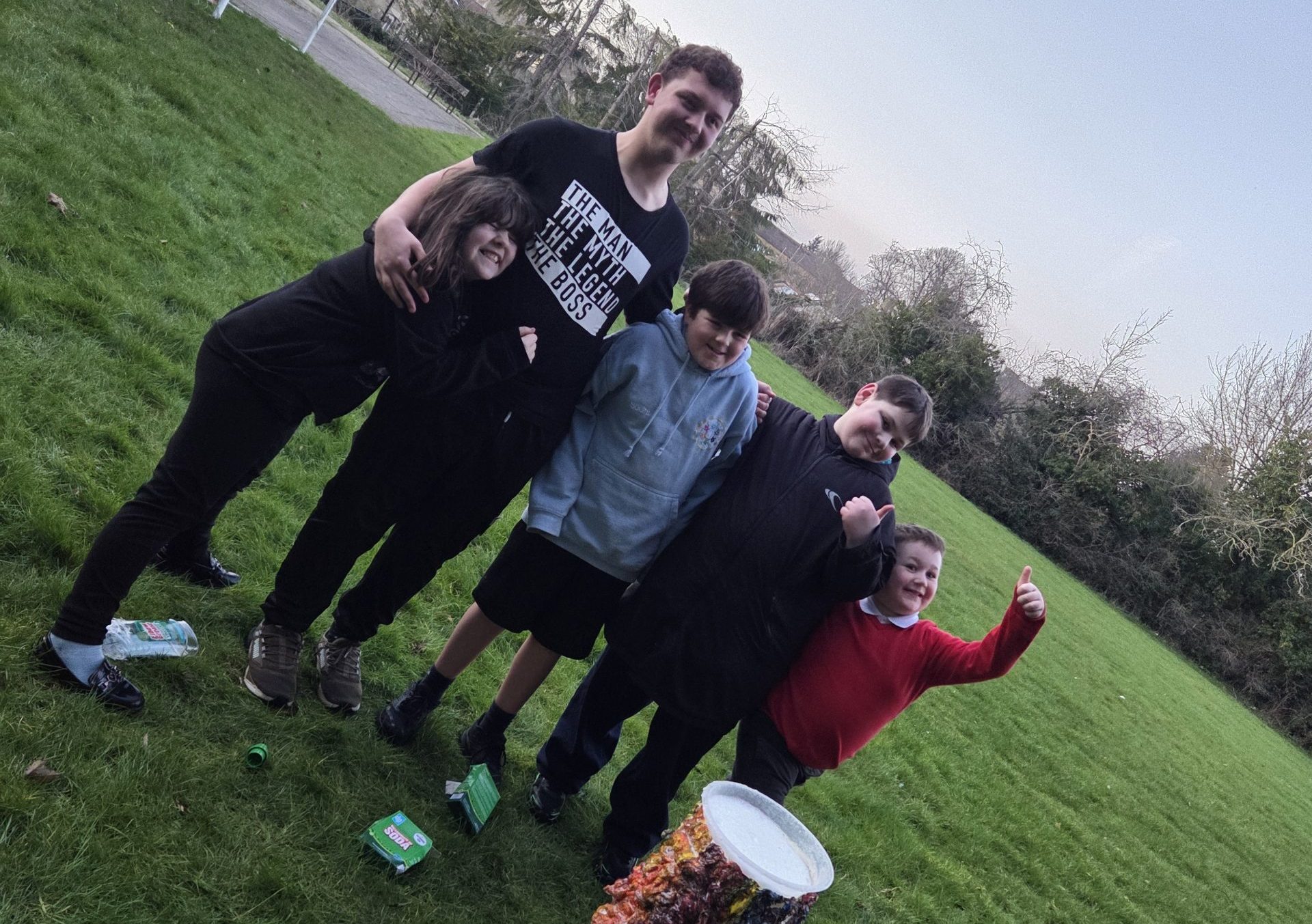 A group of children and young people stand together smiling on a grassy field beside a colourful volcano model activity. One child gives a thumbs up, with a football goal and buildings visible in the background.