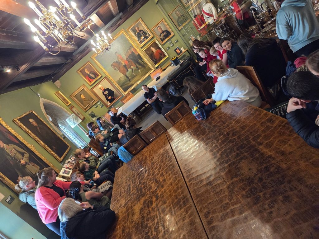 A group of children, young people and adults seated around a large wooden table during an activity in a historic museum room, surrounded by framed military portraits, display cases, chandeliers and traditional furnishings.