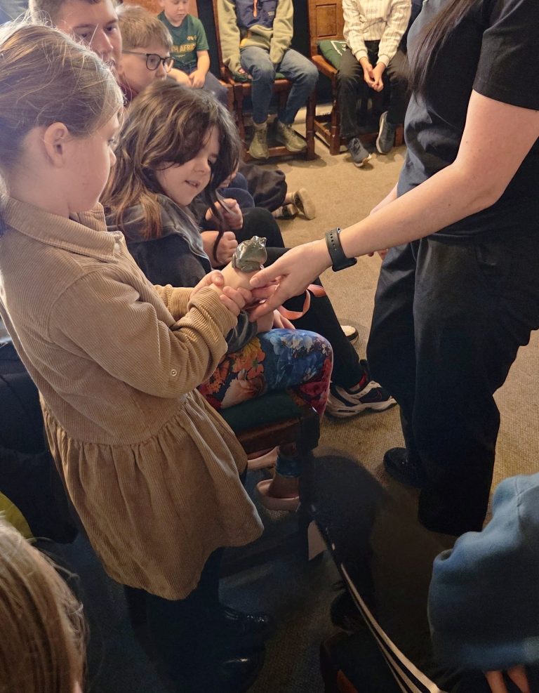 Children sit together during a hands-on group activity while an adult shows them a small object. One child carefully holds the object as others look on, creating an engaged learning moment in a community or museum-style setting.