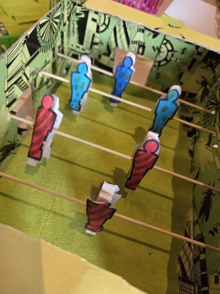 A handmade tabletop football game made from a decorated cardboard box, wooden rods and paper player figures. The red and blue players are attached to the rods inside the box, with a green playing surface underneath.