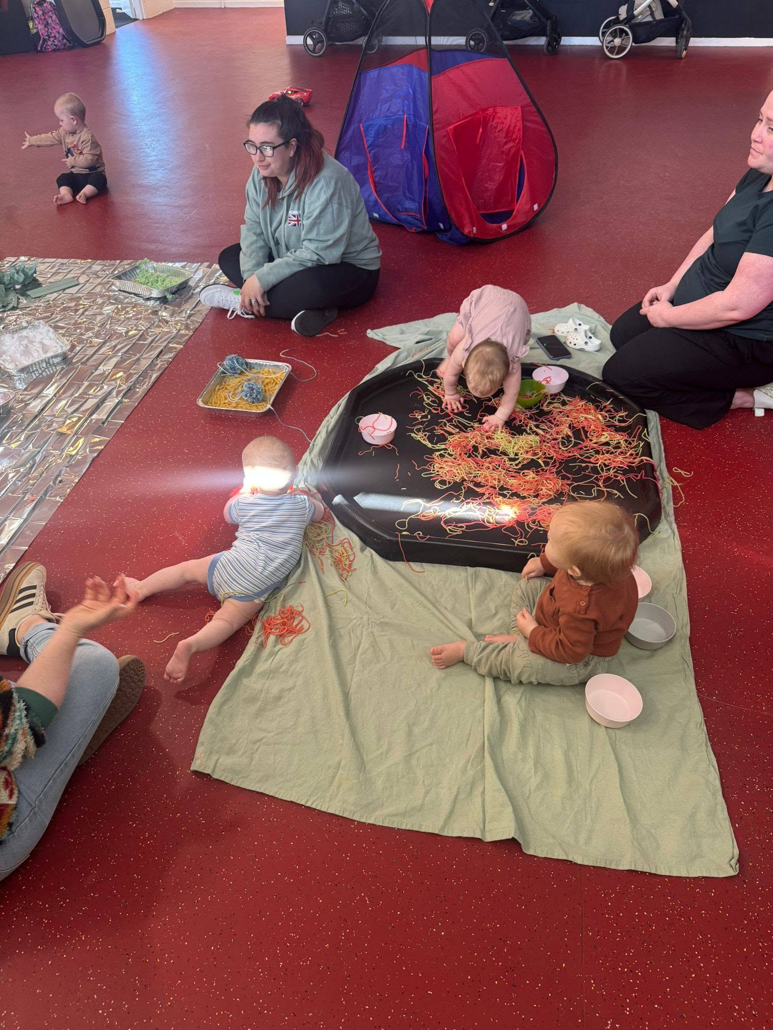 Babies and young children explore sensory play activities on a mat during a community group session. They are playing with colourful spaghetti, bowls and textured materials while adults sit nearby to supervise and support.
