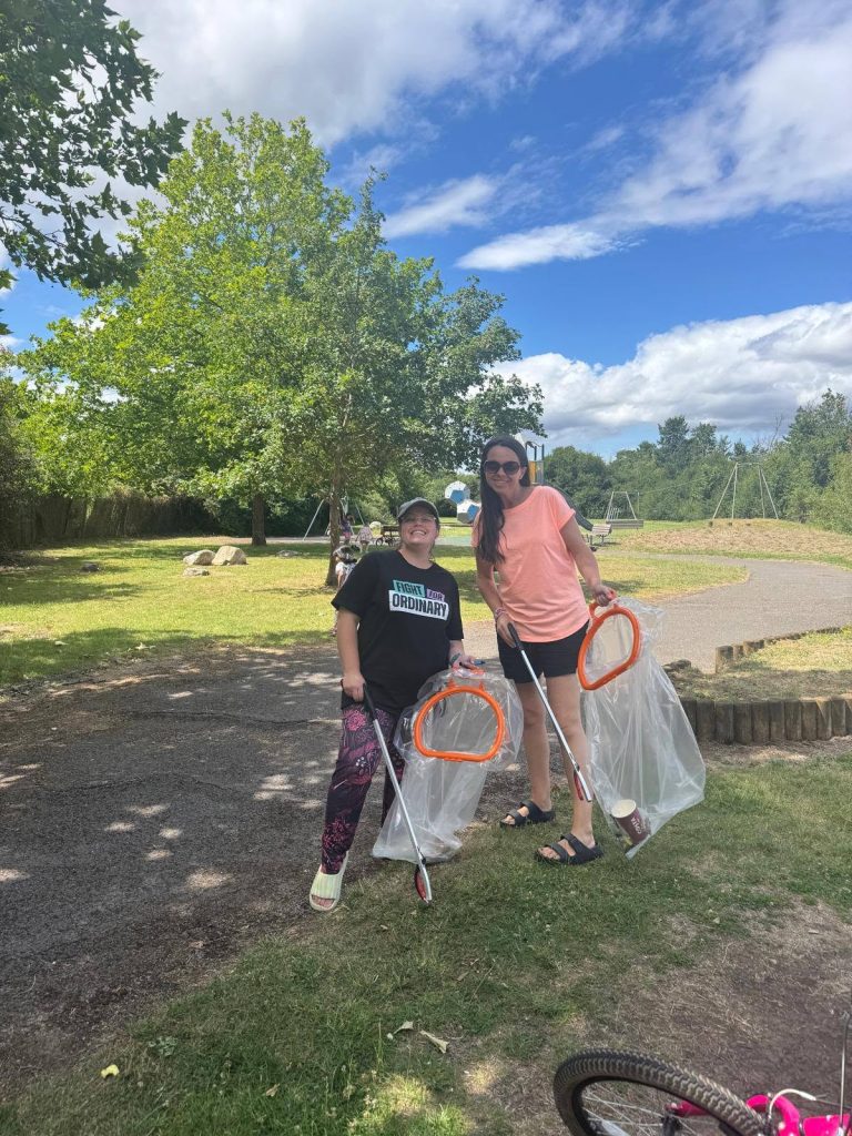 Two people smiling in a park while taking part in a litter-picking activity. They are holding litter pickers and clear rubbish bags with orange handles. Trees, grass, a play area and a bright blue sky with clouds are visible in the background.