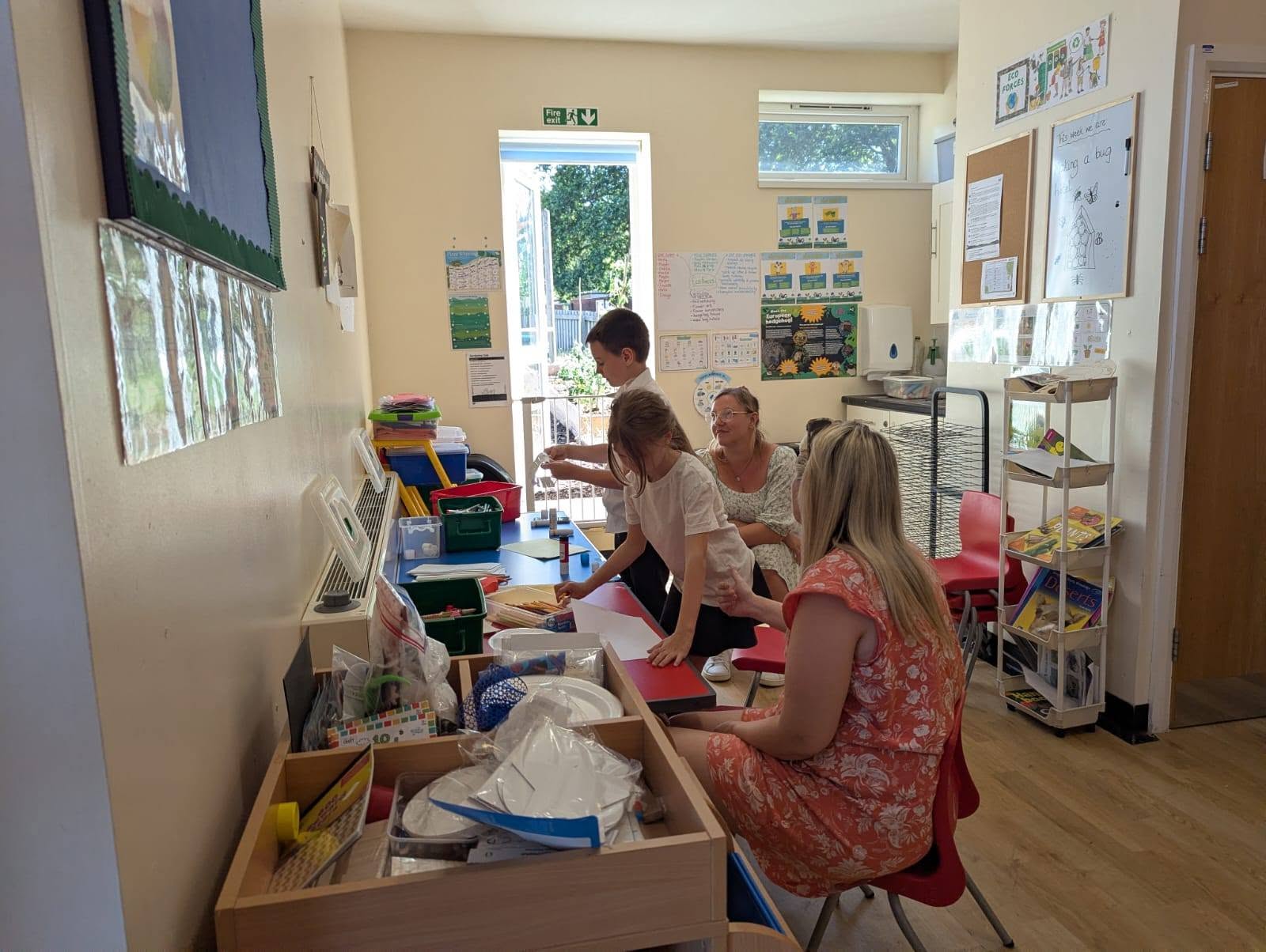 Children and adults take part in a creative activity in a bright community room. Craft materials, paper, storage boxes and learning displays are visible around the space, with adults sitting nearby to support the children.