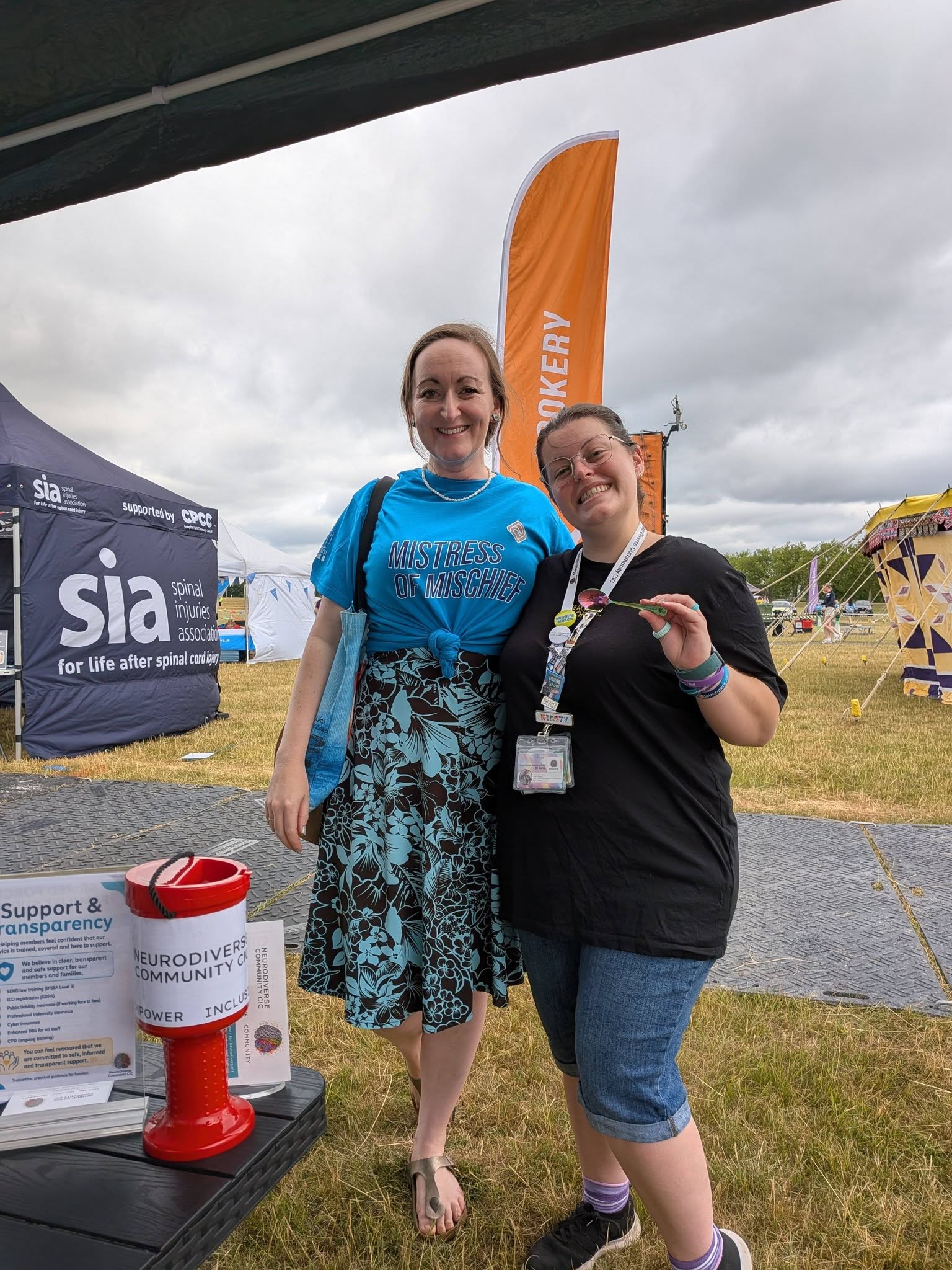 Two smiling adults standing together at an outdoor festival or community event. One is wearing a blue “Mistress of Mischief” T-shirt and patterned skirt, and the other is wearing a black T-shirt, lanyard and ID badge while holding a green spoon. A NeuroDiverse Community CIC donation bucket and information display are visible in the foreground, with event tents, an orange flag and a Spinal Injuries Association stall in the background.