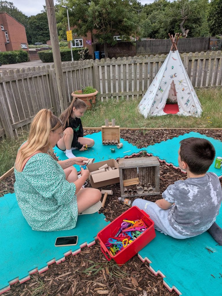 An adult and two children sitting on outdoor mats in a garden area, making bug hotels using cardboard tubes and natural materials. A wooden insect house, craft supplies, scissors and a box of materials are spread out in front of them. A small play teepee, wooden fence and greenery are visible in the background.