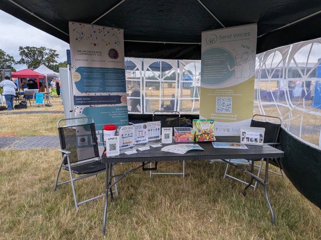 NeuroDiverse Community CIC and Send Voices information stall set up under a gazebo at an outdoor community event. A table displays leaflets, flyers, QR codes, information stands, sweets and resources. Behind the table are large banners for NeuroDiverse Community CIC and Send Voices Royal Borough of Windsor & Maidenhead. Other event stalls and people are visible in the background on a grassy field.