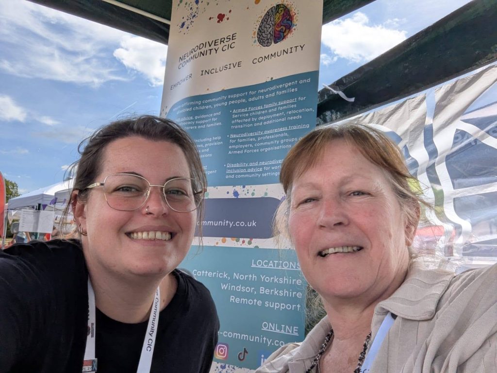 Two smiling adults taking a selfie at an outdoor community event under a gazebo. Behind them is a NeuroDiverse Community CIC banner with information about support services, locations and online contact details. Tents, a blue sky and event activity are visible in the background.
