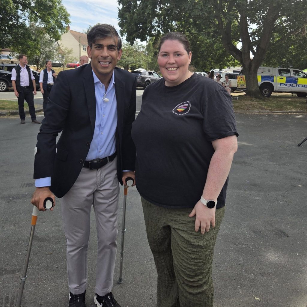 Two people stand smiling together outdoors at a community event. One person is wearing a suit jacket and using crutches, while the other is wearing a black NeuroDiverse Community CIC T-shirt. Trees, people, parked vehicles and a police van are visible in the background.