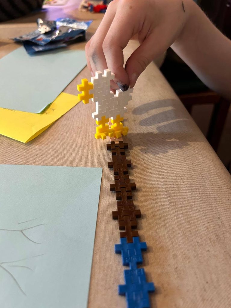 Close-up of a child’s hand holding a small pixel-style model of a white chicken with yellow feet and beak. The chicken stands above a long brown and blue block construction on a table, surrounded by coloured paper and craft materials.