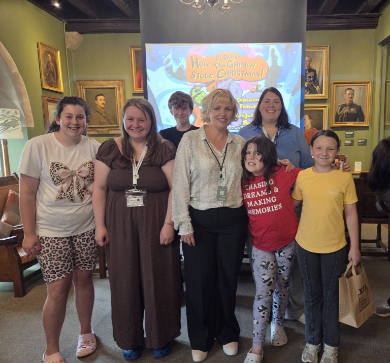 A group of adults, children and young people smiling together in a historic room decorated with framed military portraits. Behind them, a projector screen displays the menu for Dr Seuss’ How the Grinch Stole Christmas!.