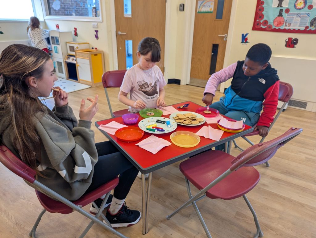 Three children sit around a table in a community room decorating biscuits with icing and sprinkles. Plates, napkins, pens and a large plate of biscuits are on the table, with play equipment and wall displays visible in the background.