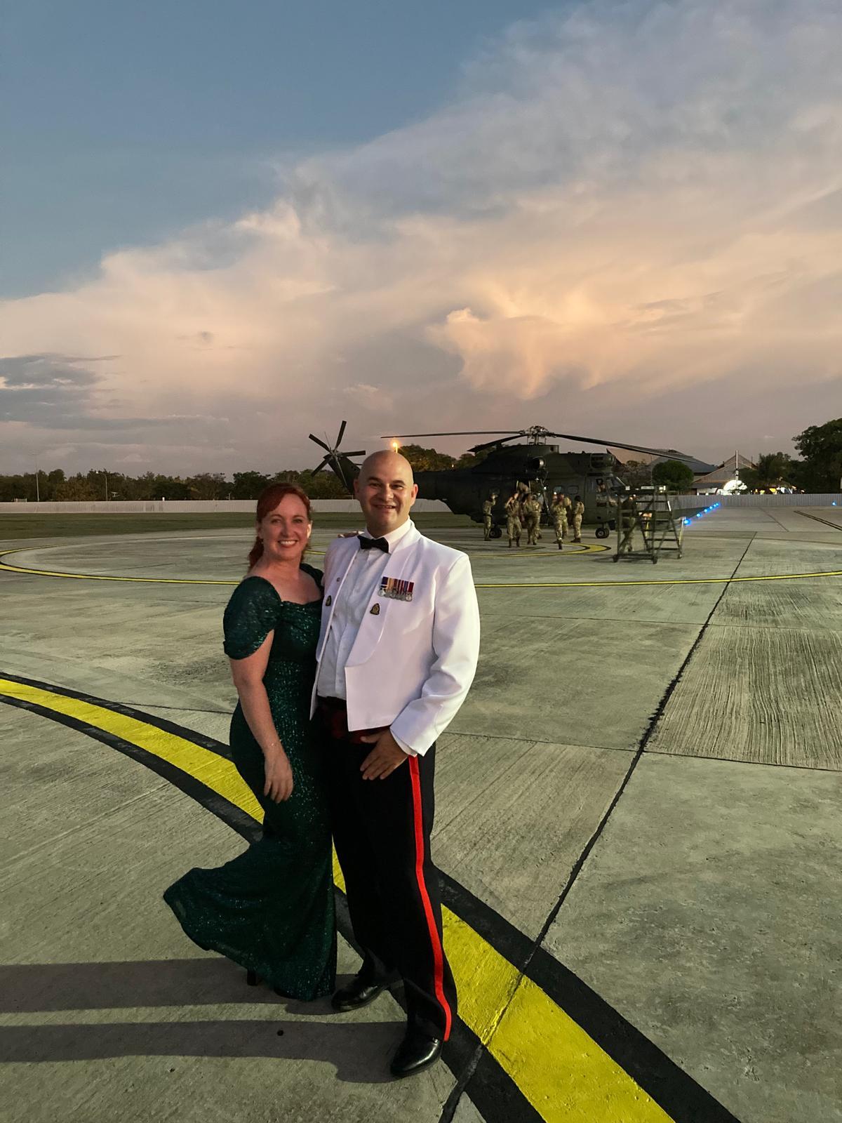 A smiling couple dressed formally at an Armed Forces event, standing on a large concrete helipad at sunset. One person is wearing a dark green evening dress, and the other is wearing a white military-style dress jacket with medals, black trousers and a bow tie. A military helicopter and personnel are visible in the background beneath a dramatic cloudy sky.