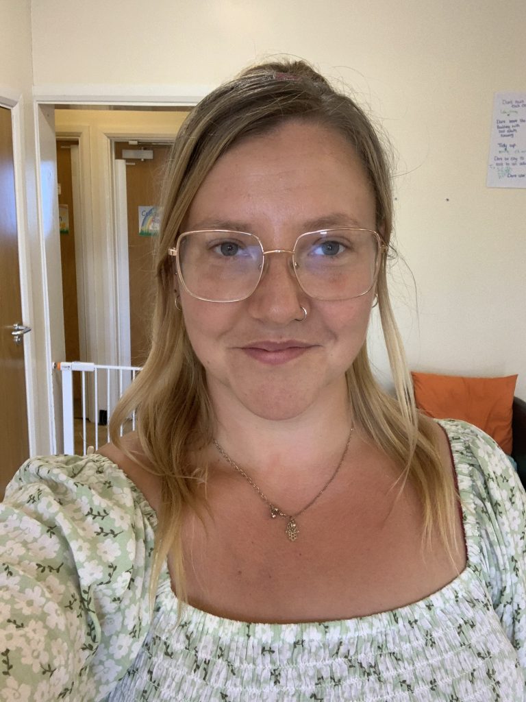 A smiling woman taking a selfie indoors. She has blonde hair, wears large glasses, a nose ring, a necklace and a pale green floral top. Behind her are open doorways, a white safety gate and an orange cushion on a chair or sofa.