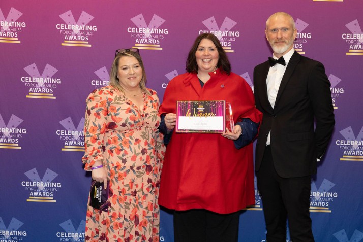 NeuroDiverse Community CIC’s Founder and CEO stands smiling with two representatives from Reed, the sponsor of the award, at the Celebrating Forces Families Awards. She is holding a winner’s certificate and trophy in front of a purple branded awards backdrop.
