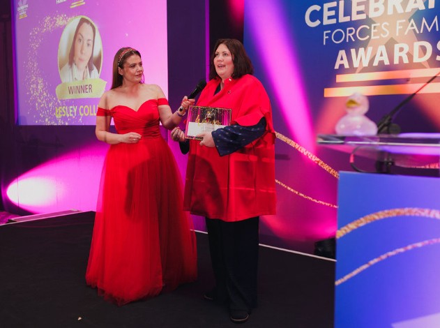 Two women stand on stage at an awards ceremony under pink and purple lighting. One woman in a red dress holds a microphone while the other, wearing a red top and black trousers, holds an award. A large screen in the background shows a winner announcement, and the stage signage reads “Celebrating Forces Families Awards.”