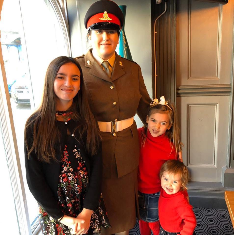 An adult in formal military uniform stands indoors with three smiling children. The children are dressed smartly, with two wearing red jumpers and the older child wearing a black floral dress and cardigan. The group are standing by a large window in a warmly lit room.
