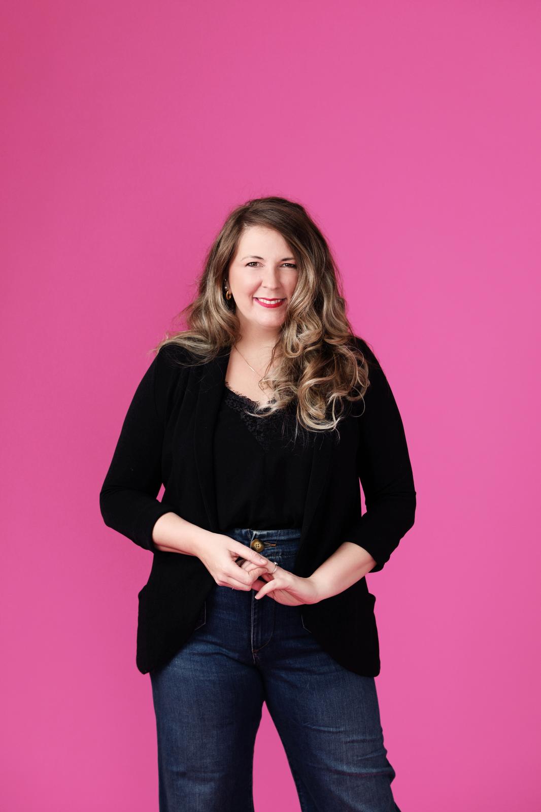 A smiling woman standing against a bright pink studio background. She has long wavy hair and is wearing a black blazer, black top and dark jeans, with her hands gently clasped in front of her.
