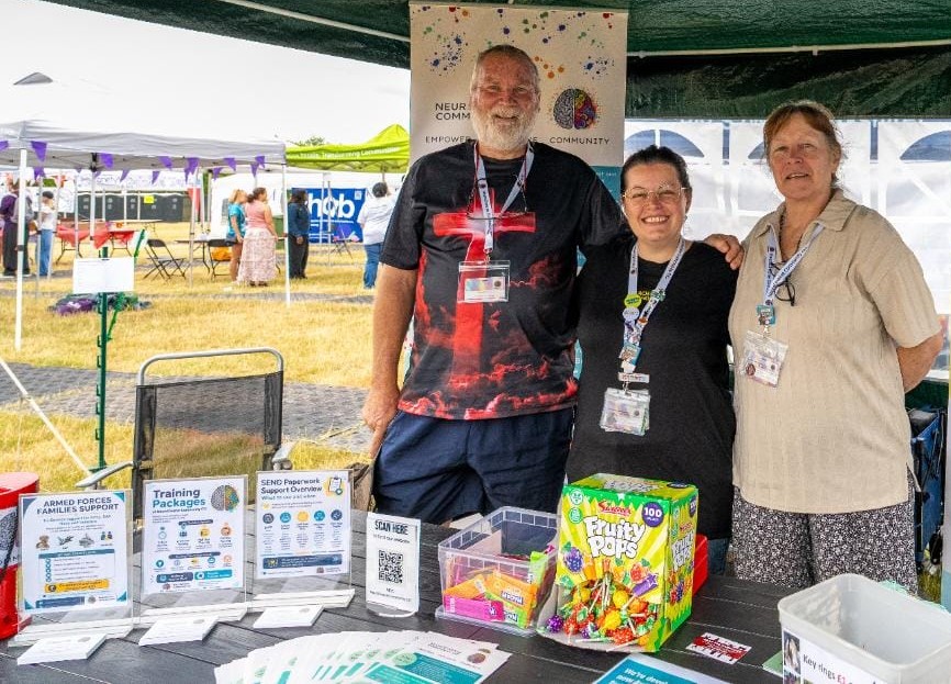 Three people stand smiling behind a NeuroDiverse Community CIC information stall at an outdoor community event. The table displays leaflets, SEND support information, Armed Forces families support materials, a sign-up QR code, sweets and resources. Gazebos and other event stalls are visible in the background.
