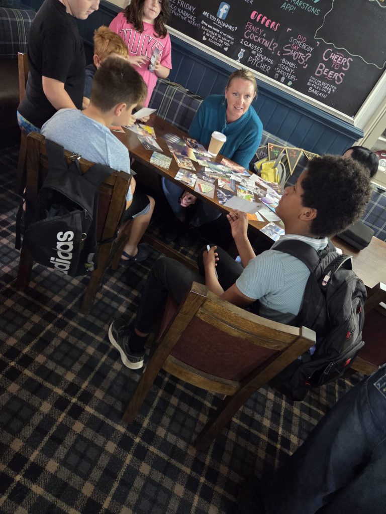 A group of young people gathered around a table with an adult facilitator, looking through and discussing a selection of printed photographs.
