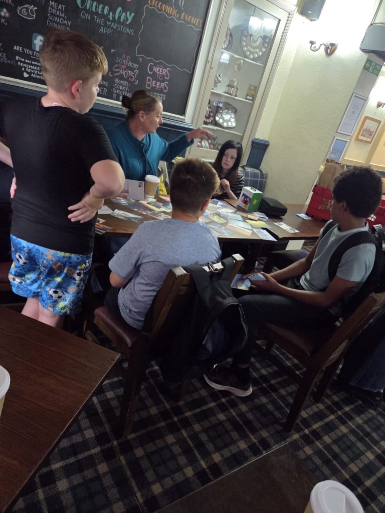 An adult facilitator points towards the photographs spread across a table while young people sit and stand around, taking part in a group discussion.