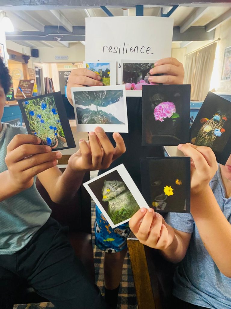 Young people hold up photographs of flowers and nature scenes in front of a handwritten sign reading “resilience”.