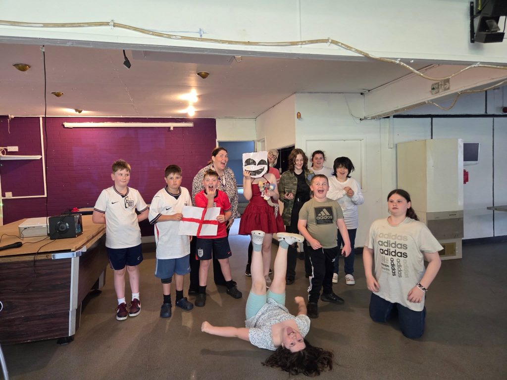 A group of children and adults pose playfully inside a community room. In the center foreground, a girl lies on her back upside down on the floor, laughing with her legs in the air. Surrounding her, group members make silly faces and pose, including a boy in a red shirt holding a drawn paper England flag, a person in a red dress holding a mask over their face, and kids in sports shirts smiling and laughing.
