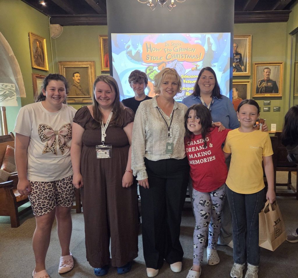 A group of children and adults pose for a photo inside a room with olive green walls lined with framed historical portraits. Behind them, a large screen displays a title slide for Dr. Seuss' How the Grinch Stole Christmas!