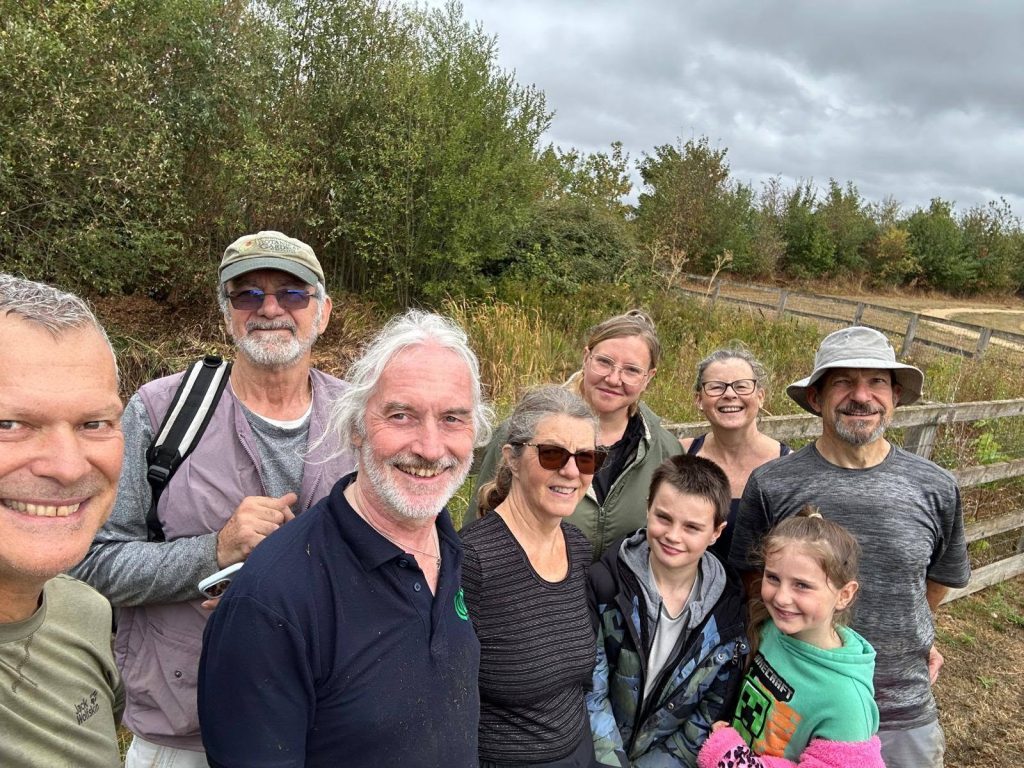 Group selfie outdoors at a nature reserve, showing seven adults and two children smiling together beside trees, wild vegetation and a wooden fence under a cloudy sky.