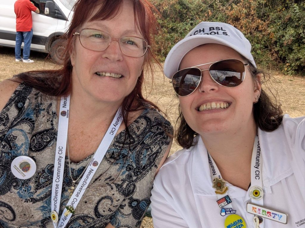 Two NeuroDiverse Community CIC team members smiling for a selfie at Rock the Rec. Both wear NDC-branded lanyards and badges, with one wearing sunglasses and a BSL-themed cap and several inclusion and wellbeing badges.