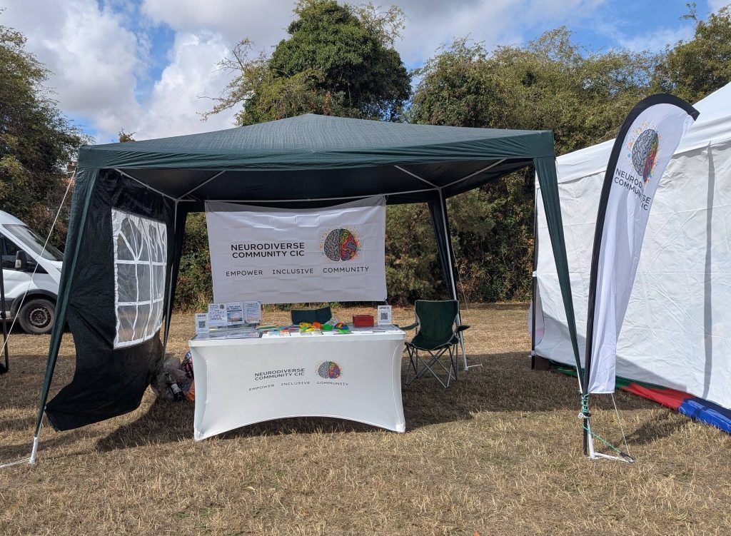 NeuroDiverse Community CIC information stand at Rock the Rec, with a green gazebo, branded banner, tablecloth and feather flag. The table displays information, resources and colourful sensory items, set up outdoors on the event field.