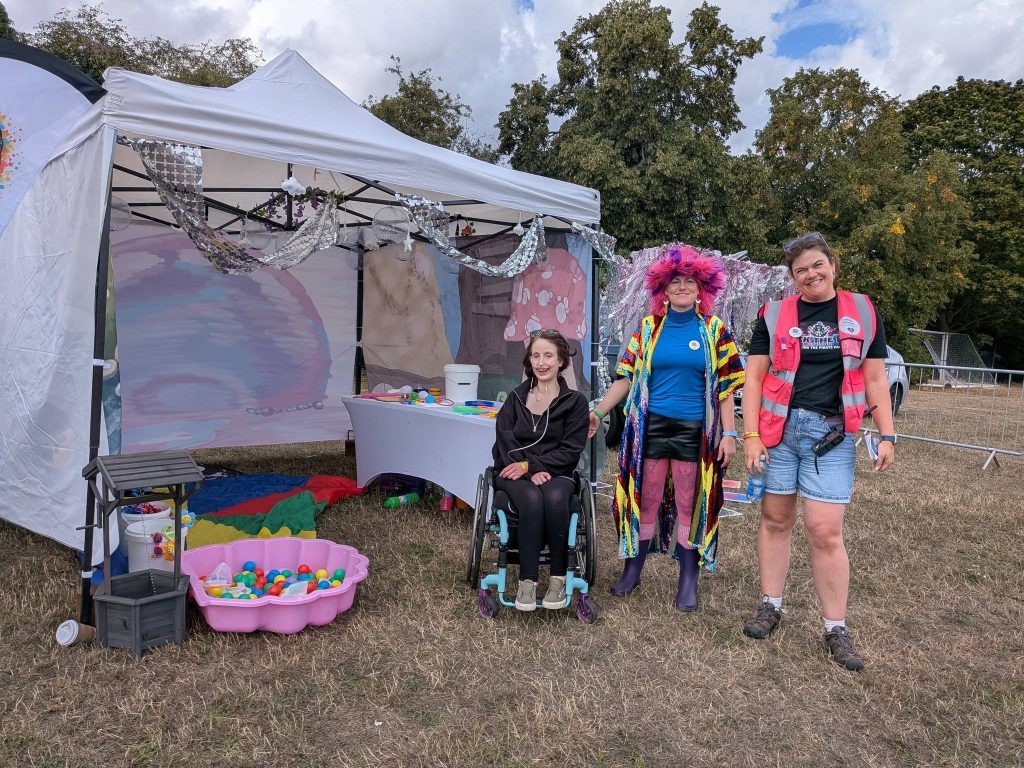 Three women smiling beside a sensory activity tent at an outdoor community event. One woman is seated in a wheelchair, another wears a colourful outfit and bright pink-and-purple wig, and the third wears a high-visibility event vest. Sensory toys, a ball tray and decorations are displayed around the tent.