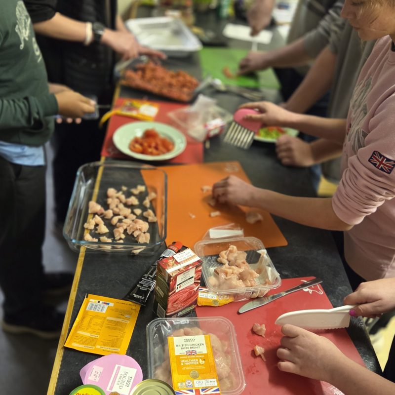 **Alternative text:** A group of young people and adults preparing food together in a kitchen. They are chopping chicken and vegetables on coloured chopping boards, with ingredients such as tomatoes, beans, sauce and sour cream laid out on the counter.