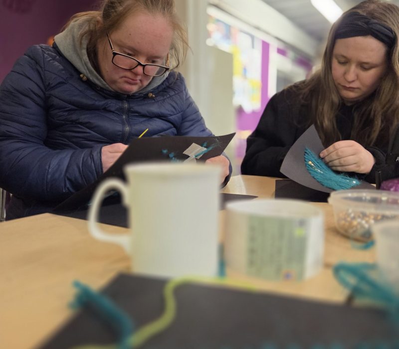 Two women sit at a table taking part in a craft activity, carefully working with coloured paper and materials. A mug, craft supplies and small colourful items are visible in the foreground.
