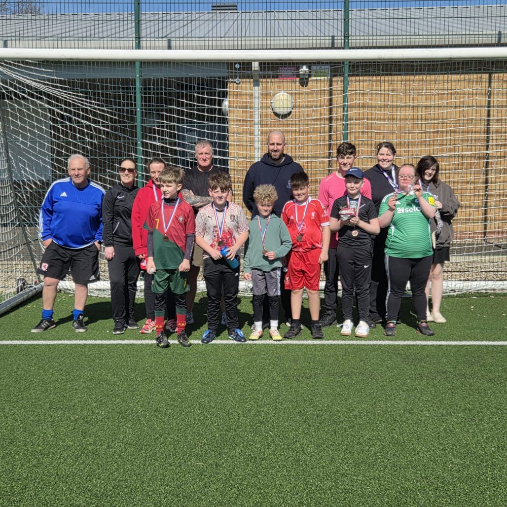 A group of adults and children standing together in front of a football goal on an outdoor artificial grass pitch. Several children are wearing football kits and medals, with some holding trophies, on a sunny day.