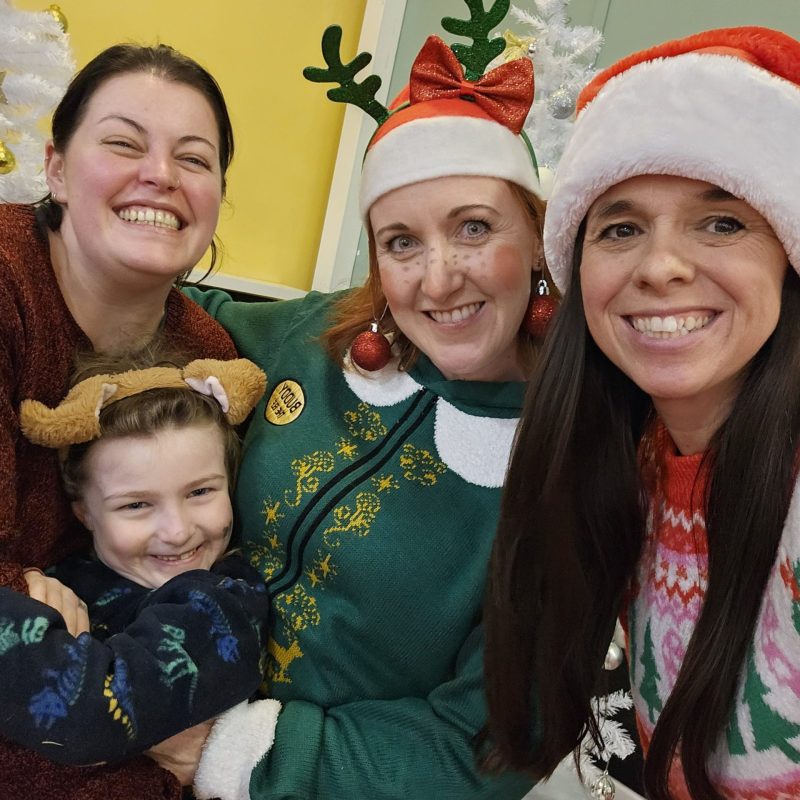 **Alternative text:** Three adults and a child smiling together for a festive photo. They are wearing Christmas jumpers, Santa hats and reindeer accessories, with white Christmas trees and decorations in the background.